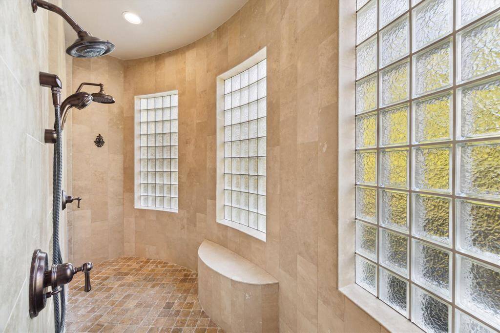 Spacious walk-in shower in the primary bathroom featuring a dual shower head system, beige stone tile walls, a herringbone-patterned tile floor, and privacy glass block windows that let in natural light.