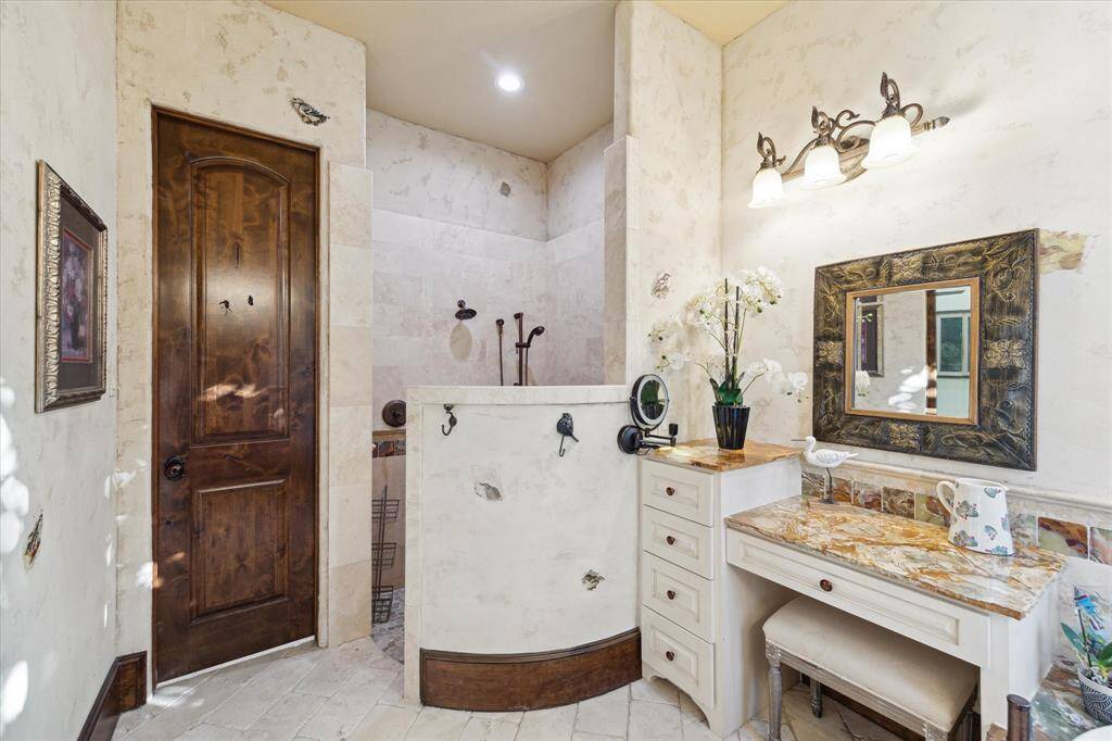 This is another secondary appointed bathroom featuring a custom curved vanity with upscale countertops, ornate mirror, and a mix of natural stone tiles. There's a built-in makeup desk and the room is well-lit with a combination of natural and artificial light.