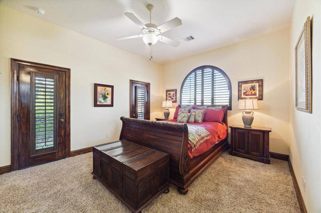 This well-lit secondary bedroom on the second floor featuring a large arched window with plantation shutters, a ceiling fan, plush carpeting, and elegant dark wood furnishings.