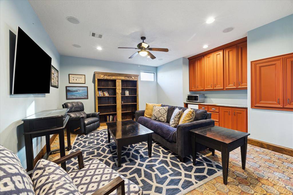 The room features a cozy entertainment area with plush seating, a wall-mounted flat-screen TV, and a bookshelf. There's also a small kitchenette with ample cabinet space and a microwave. The space is finished with patterned area rugs and has a ceiling fan for comfort.