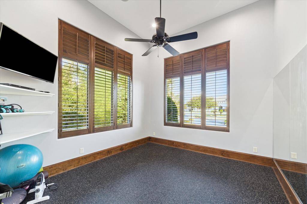 Well-lit corner room featuring two large windows with wooden blinds, a ceiling fan, and a durable, dark speckled floor. There are built-in shelves on one wall, and the space currently holds exercise equipment, suggesting it could be used as a home gym or a multi-purpose room.