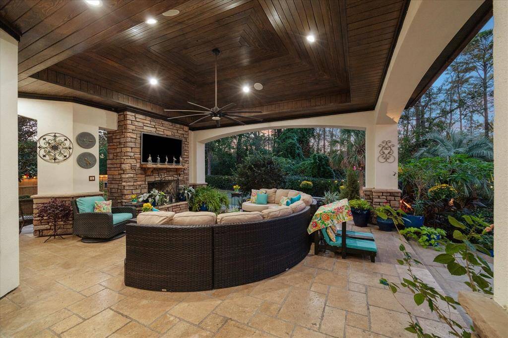 Inviting covered outdoor living space featuring a cozy seating area with plush cushions, a stone fireplace for ambiance and warmth, a rich wooden ceiling with recessed lighting and a ceiling fan, all surrounded by lush greenery providing a serene backdrop.