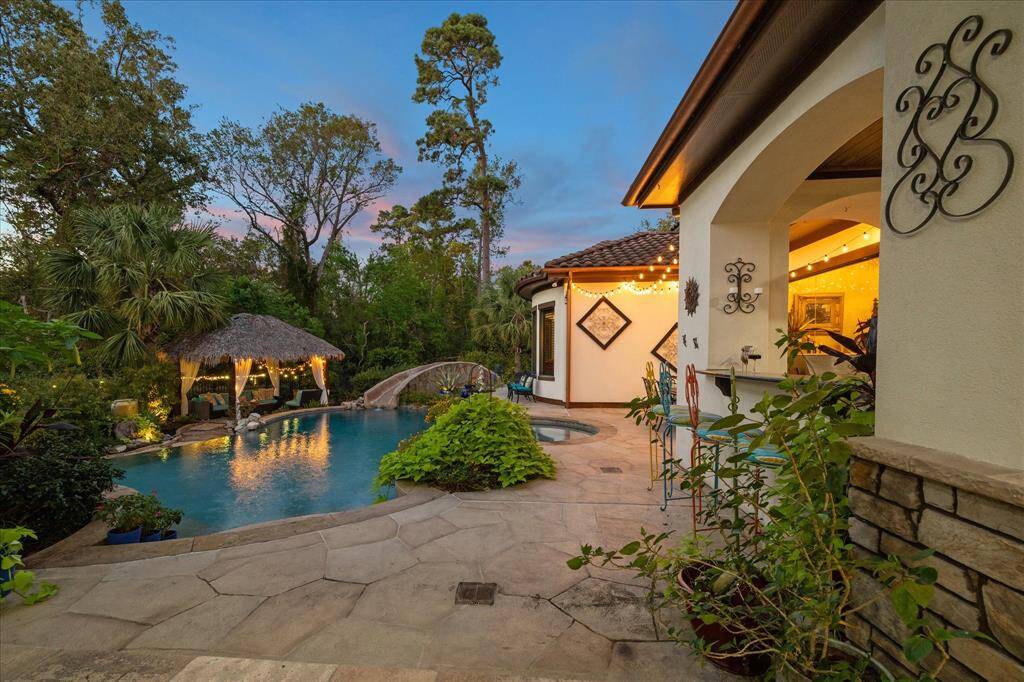 Exquisite poolside view showcasing a tropical-style swimming pool with a charming thatched-roof hut and stone bridge, surrounded by lush greenery. The home features a warm, inviting exterior with elegant decorative ironwork and ambient lighting, perfect for evening entertainment.