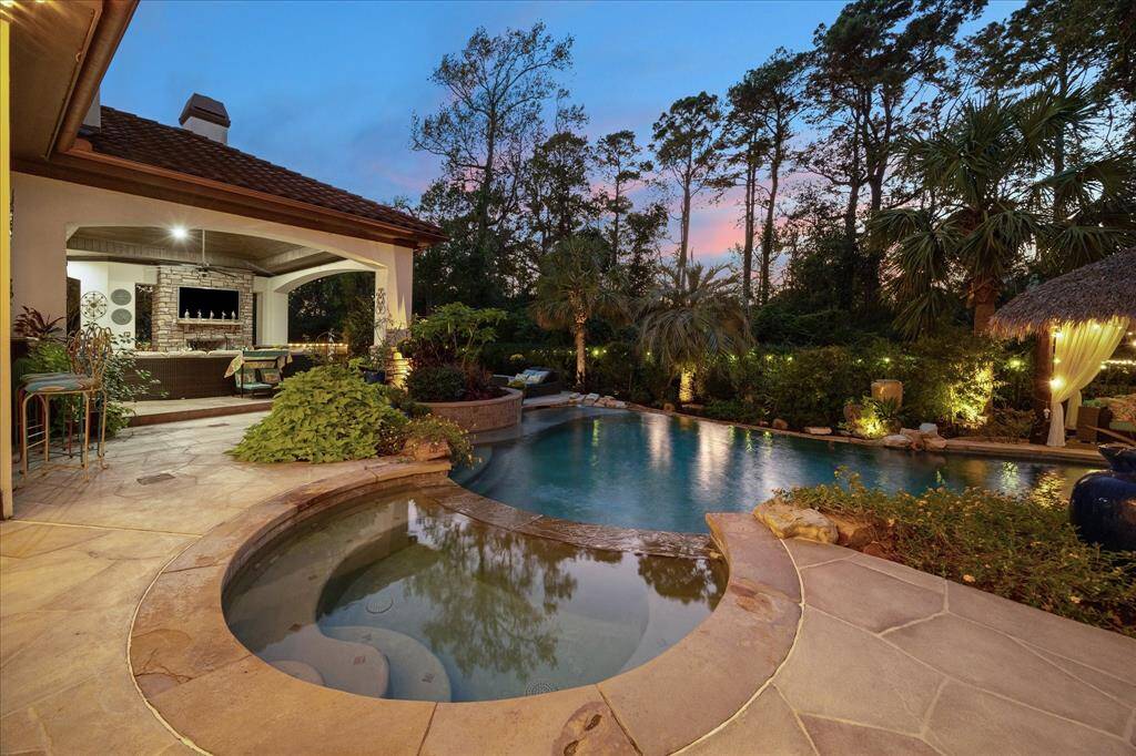 This is another luxurious outdoor living space featuring an elegant pool with a spa, surrounded by mature trees. There's a covered outdoor kitchen with a bar and a grill, perfect for entertaining. The area is well-lit, showcasing a beautiful sunset, suggesting a serene and private atmosphere.