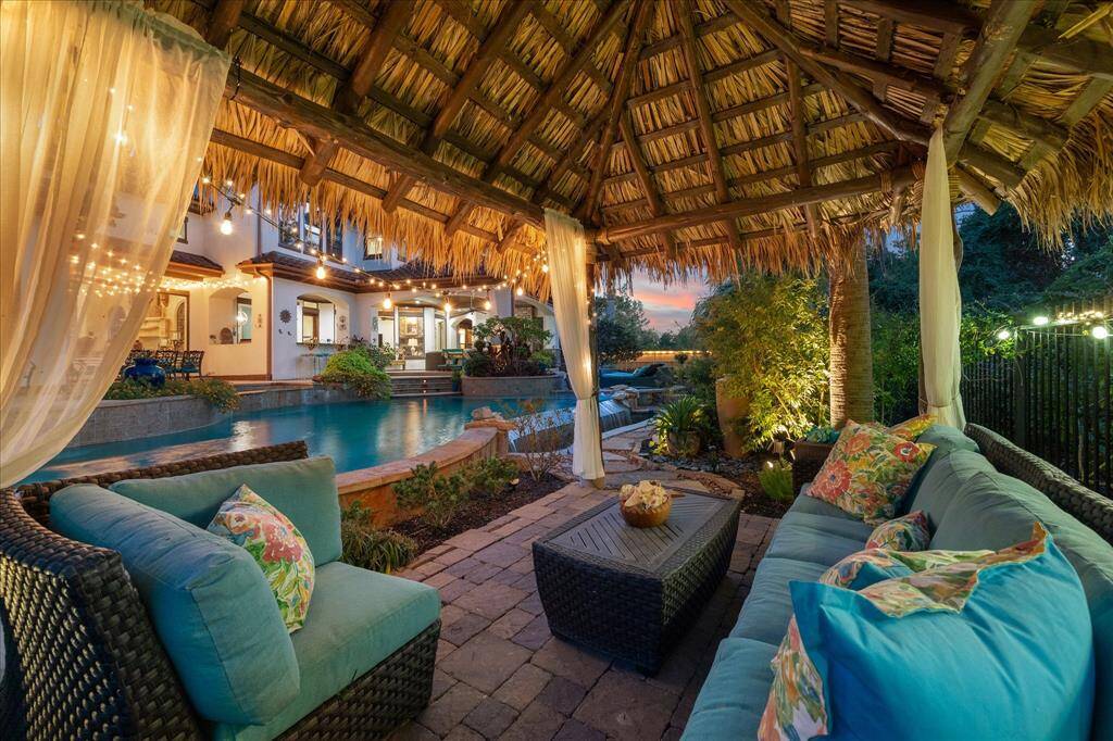 This image showcases a tropical-inspired outdoor living space with a thatched roof gazebo, comfortable seating, and a view of a serene pool, all set against the backdrop of a home with warm lighting and a sunset sky.