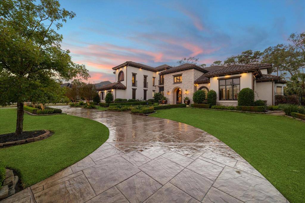 This is a luxurious two-story estate featuring a Mediterranean architectural style with a well-manicured lawn, elegant landscaping, and a stamped concrete driveway, captured at dusk under a beautifully colored sky.