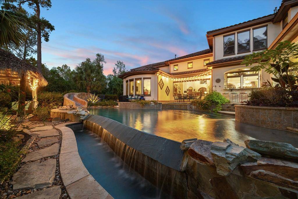 A view of the resort-style pool with a water slide and waterfall, set against a backdrop of lush landscaping and a stunning sunset sky. Outdoor lighting enhances the ambiance, perfect for evening entertainment.