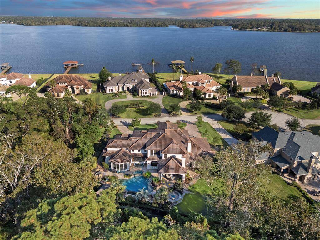 Aerial view of a luxurious lakeside property featuring a large house with a tile roof and a well-manicured lawn. The residence has a private swimming pool and is situated within a community of similarly upscale homes, offering a picturesque view of the water and a serene living environment.