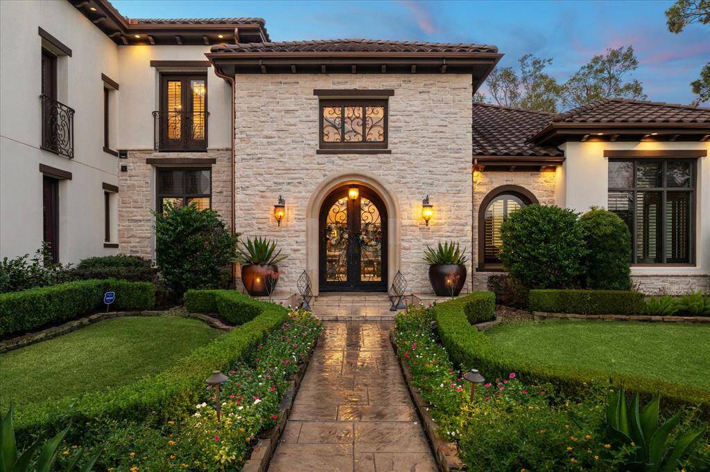 This elegant 6 bedroom Mediterranean style home features a stone facade and a grand arched entryway. The exterior is illuminated by warm lighting, enhancing the home's inviting ambiance.