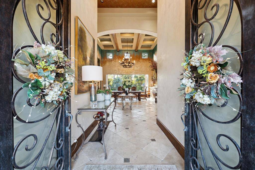 You're looking at an elegant entryway with decorative wrought-iron doors adorned with floral wreaths. Beyond lies a spacious interior with tiled flooring and a high, beamed ceiling, providing a welcoming and upscale atmosphere.