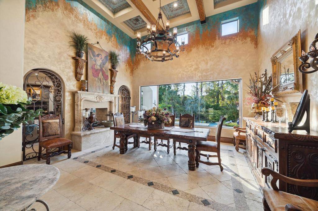This is an elegant dining room exuding warmth and charm, featuring a stone fireplace, 20 ft  beamed ceilings, and a large window overlooking the pool area. The room is adorned with decorative wrought-iron elements, creating an inviting atmosphere, ideal for entertaining friends and family..