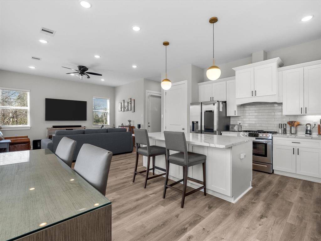 This photo showcases a modern, open-concept kitchen and living area. The kitchen features white cabinetry, stainless steel appliances, and a central island with seating. The living area has a ceiling fan, a wall-mounted TV, and ample natural light from the windows, complemented by wood flooring throughout.