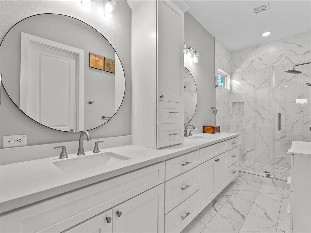 This bathroom features a modern design with dual sinks, a large circular mirror, and sleek white cabinetry. It includes a spacious glass-enclosed shower with elegant marble-patterned tiles, offering a bright and contemporary feel.