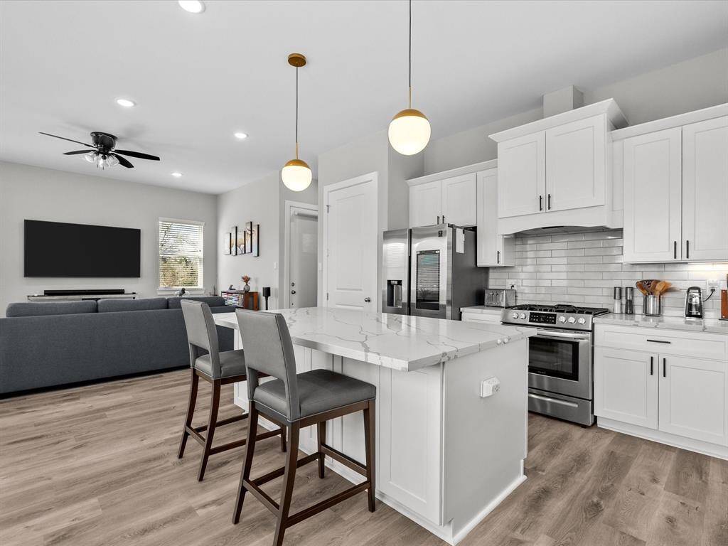 This photo showcases a modern, open-concept kitchen and living area. The kitchen features white cabinetry, stainless steel appliances, and a marble island with seating. The living space includes a cozy seating area, a large TV, and ample natural light from a window. The room has a clean, contemporary design with wood flooring and pendant lighting.