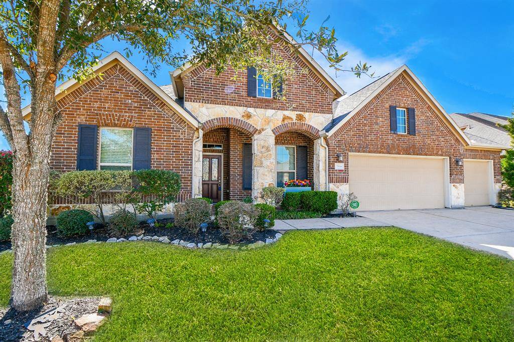 Charming brick home with a well-maintained lawn, featuring a welcoming stone archway entrance. The property includes a spacious three-car garage, decorative shrubs, and mature trees, offering great curb appeal.