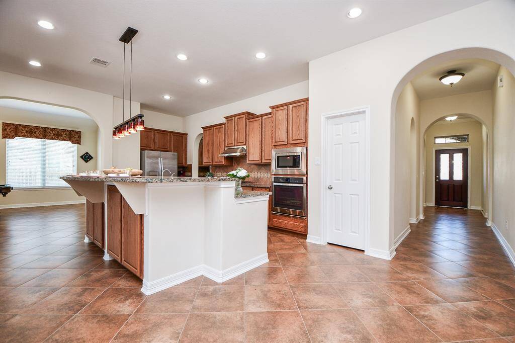 This spacious kitchen features elegant wood cabinetry, granite countertops, and modern stainless steel appliances. It opens to a large, well-lit dining area with tile flooring throughout. The hallway offers a welcoming entry with arched doorways for a stylish touch.