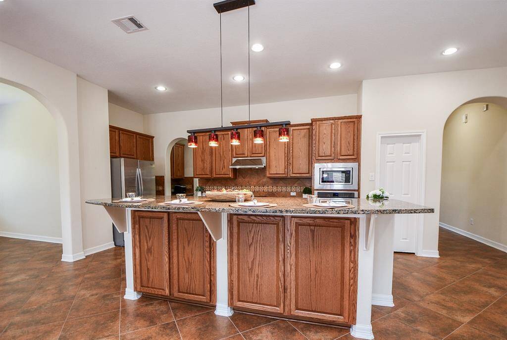 This kitchen features warm wood cabinetry, granite countertops, and a central island with stylish pendant lighting. It includes stainless steel appliances, a built-in microwave, and tile flooring. Arched doorways add an elegant touch, enhancing the open and inviting space.