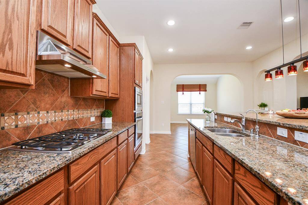 This kitchen features granite countertops, wooden cabinetry, and a modern tile backsplash. It includes stainless steel appliances, a gas cooktop, and a double sink. The space is open and well-lit with recessed lighting, leading into a bright dining area.