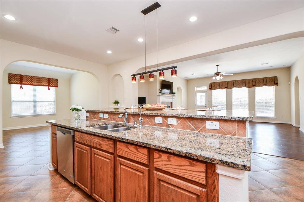 This spacious open-concept kitchen and living area features a large granite island, stylish pendant lighting, and tile flooring. The room is bright with natural light from multiple windows and offers a cozy living space with a ceiling fan and fireplace.