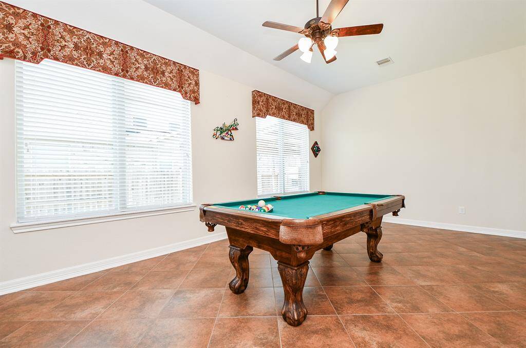 This spacious game room features tile flooring, and large windows with decorative valances, providing plenty of natural light. A ceiling fan adds comfort to the airy, welcoming space.