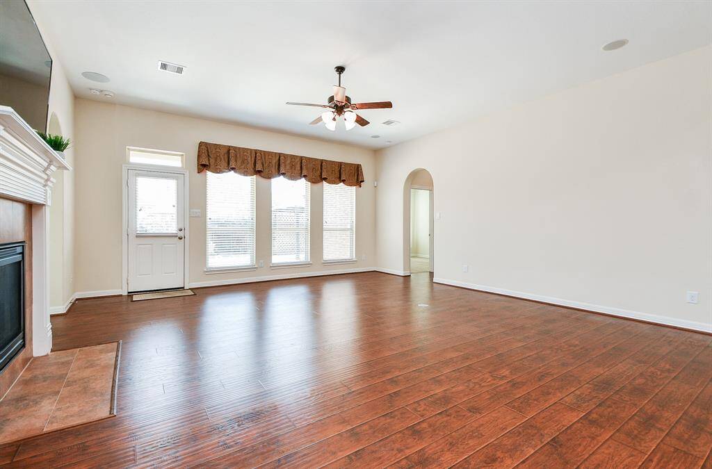 This spacious living room features hardwood floors, a cozy fireplace, large windows with ample natural light, and a ceiling fan. The neutral walls provide a versatile backdrop, and there’s a convenient entryway door and an adjoining archway leading to another room.