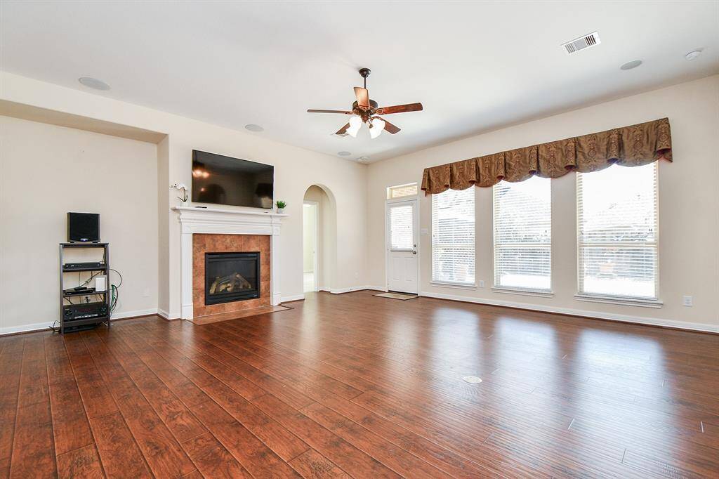This spacious living room features hardwood floors, a cozy fireplace with a TV mounted above, and large windows that provide ample natural light. A ceiling fan enhances the comfort of the room, and there's an archway leading to another area of the home.