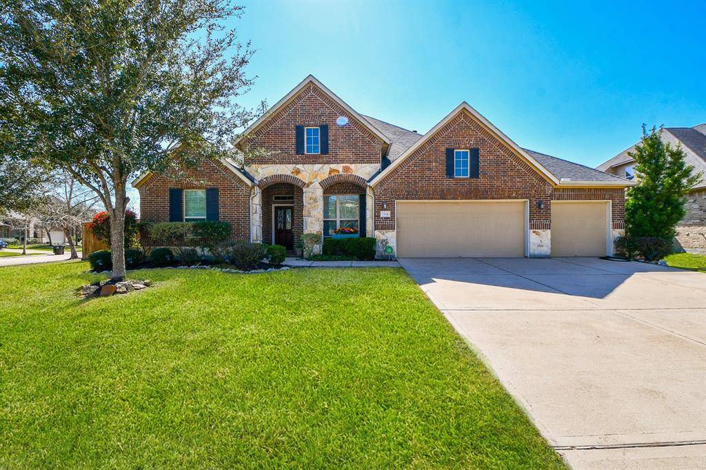 This is a charming brick home with a well-maintained lawn and mature trees. It features a three-car garage and a welcoming front entrance with stone accents. The property sits on a spacious corner lot in a suburban neighborhood.