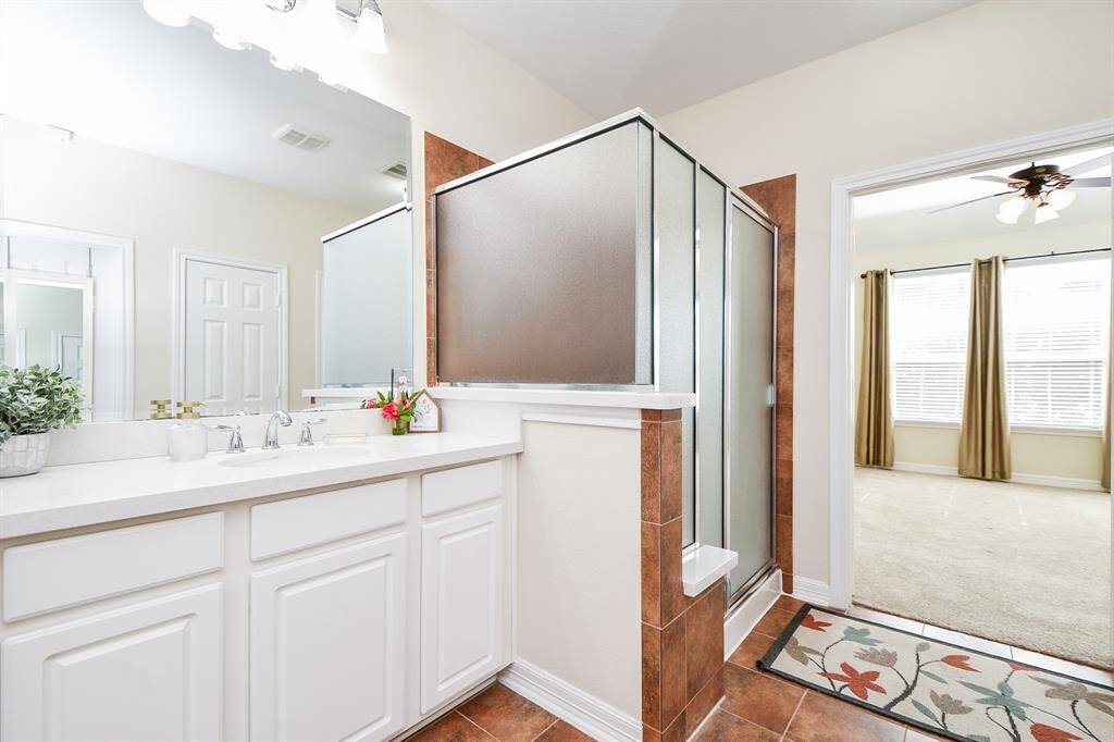 This bright bathroom features a dual-sink vanity with white cabinetry, a frosted glass shower, and warm tile accents. It opens into a carpeted room with large windows and ceiling fan, providing a seamless flow between spaces.