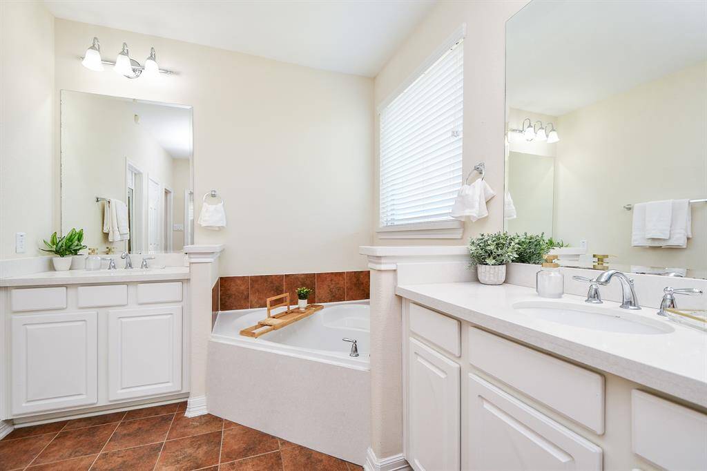 This bathroom features a bright, spacious design with dual sinks, a large mirror, and elegant fixtures. It includes a cozy corner bathtub with surrounding tiles, ample counter space, and natural light from a window with blinds.