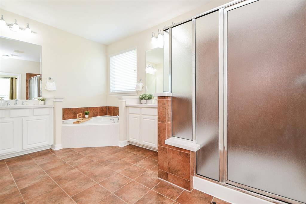 This spacious bathroom features a large walk-in shower with frosted glass, a corner bathtub, dual sinks with ample counter space, and warm-toned tile flooring. Natural light enhances the bright, clean atmosphere.