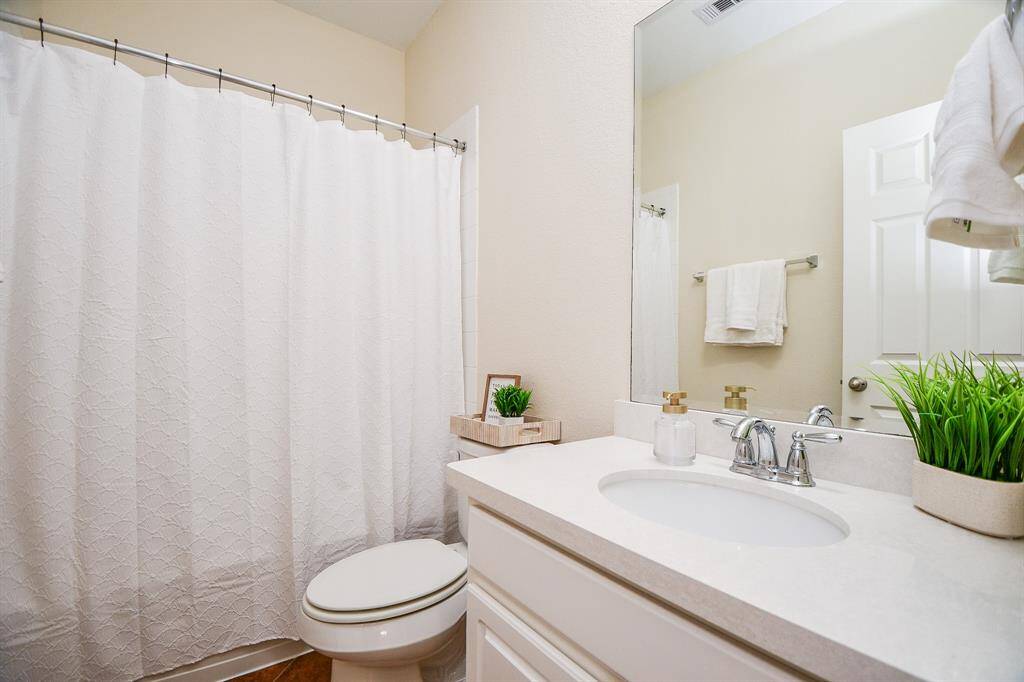 This bathroom features a clean, modern design with a white vanity and countertop, a sleek faucet, and a large mirror. It includes a shower with a white curtain, neutral walls, and decorative touches like potted plants and towels.