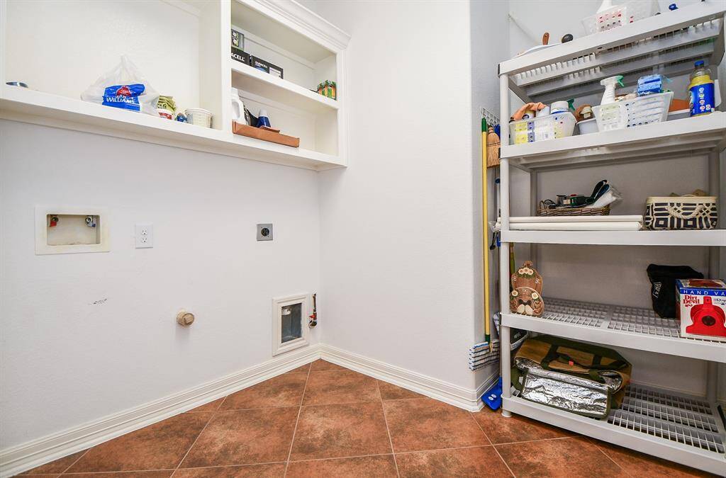 This photo features a laundry room with white walls, built-in shelving, and a tiled floor. It includes hookups for a washer and dryer, additional storage shelves, and ample space for organizing cleaning supplies.