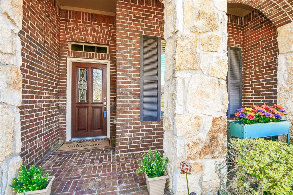 This inviting entrance features a rich wooden door with decorative glass panels, set against a charming brick exterior. The porch is accented with stone columns, shutters, and colorful flowers, creating a warm, welcoming atmosphere.