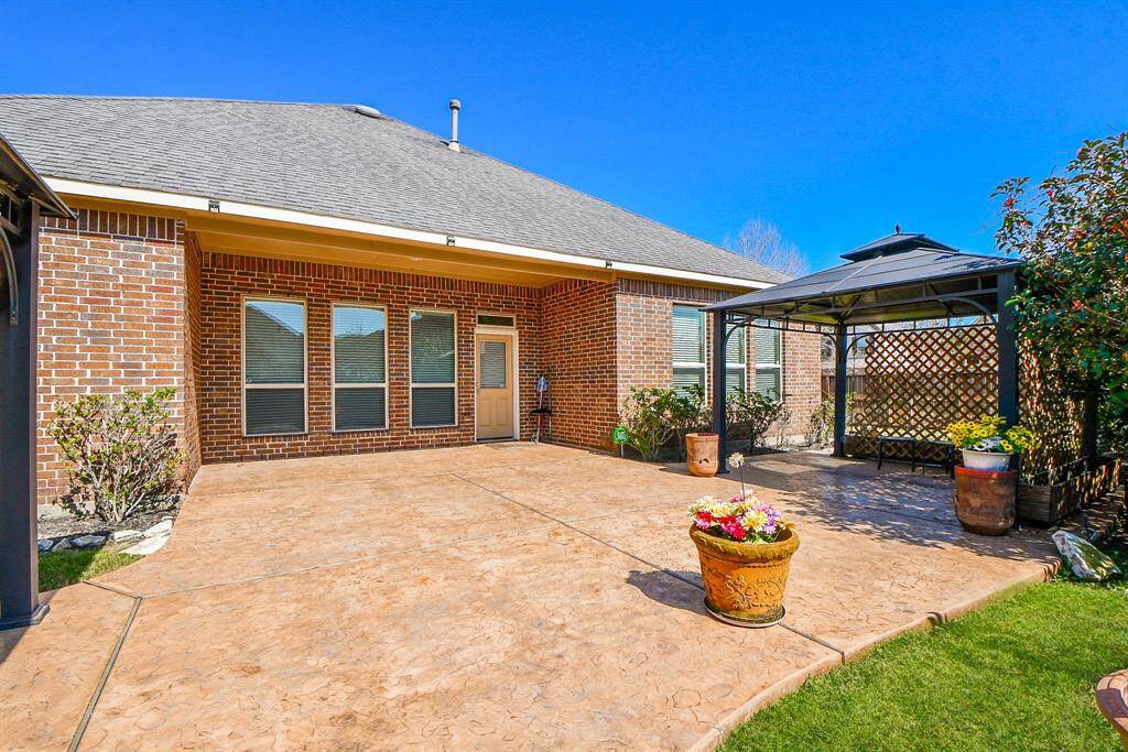 This photo showcases a spacious, well-maintained backyard patio with a brick exterior. It features a covered gazebo area, perfect for outdoor entertaining, and is surrounded by neatly landscaped greenery.