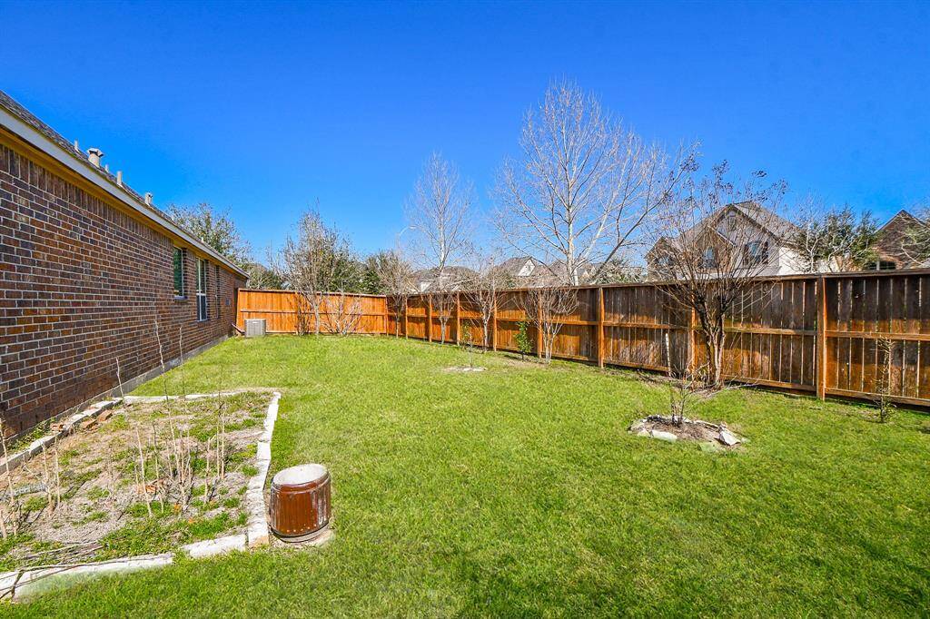 This photo showcases a spacious backyard with a well-maintained lawn, bordered by a wooden fence for privacy. The side of a brick house is visible, and there are a few young trees and a small garden area, ideal for outdoor activities or gardening.