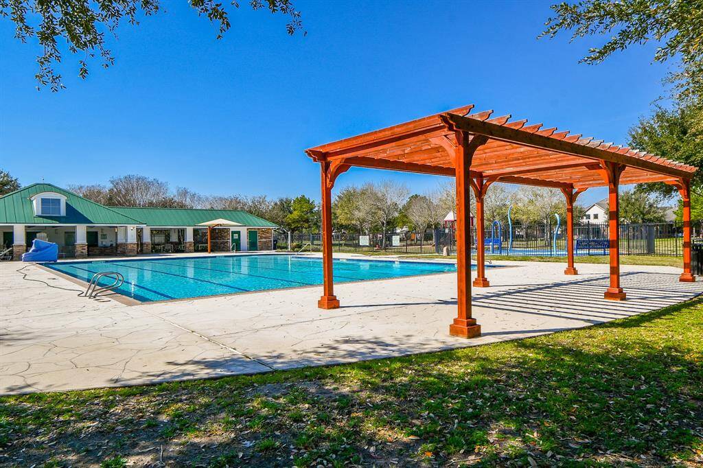 This photo showcases a serene neighborhood pond with a fountain, surrounded by well-maintained grass and houses in the background, under a clear blue sky. Ideal for a peaceful living environment.