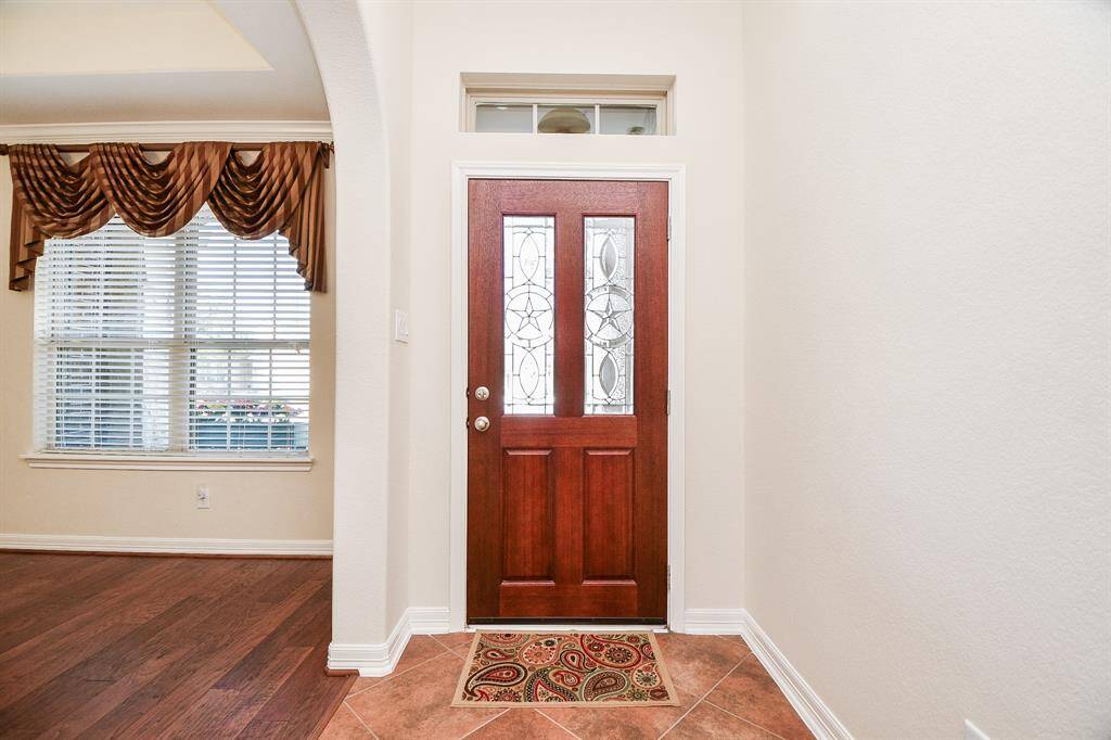This photo showcases a welcoming entryway with a rich wooden door featuring decorative glass panels. Adjacent is a room with large windows dressed with elegant drapes, and a contrast of tiled and hardwood flooring adds warmth and character.