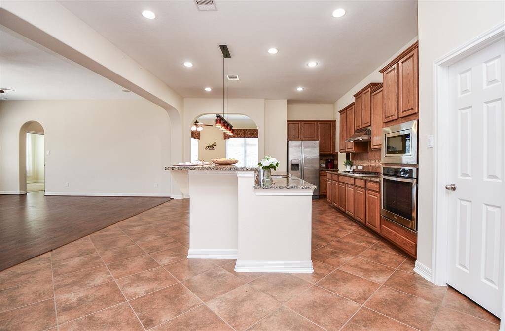This photo showcases a spacious kitchen with granite countertops, wooden cabinetry, and stainless steel appliances, including a double oven and a large refrigerator. The open layout connects to a living area with hardwood flooring, creating a seamless flow ideal for entertaining. Recessed lighting and pendant lights illuminate the space, giving it a modern and inviting feel.