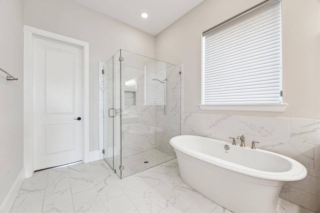 A luxurious primary bathroom with a freestanding soaking tub positioned beneath a large window, allowing for soft natural light. The glass-enclosed walk-in shower features elegant marble-inspired tile, creating a clean and sophisticated look.