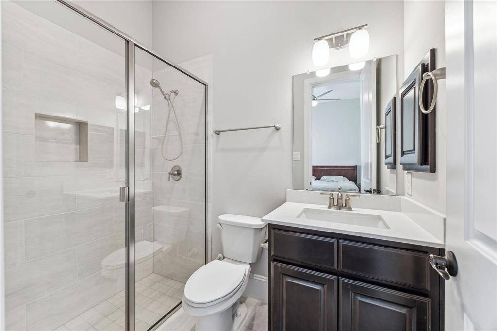 This ensuite bathroom offers a sleek glass-enclosed walk-in shower with built-in shelving for added functionality. A quartz countertop vanity with dark wood cabinetry provides ample storage, while brushed nickel fixtures complement the neutral color scheme. Connected directly to the first-floor secondary bedroom, this space ensures privacy and convenience for guests or residents.