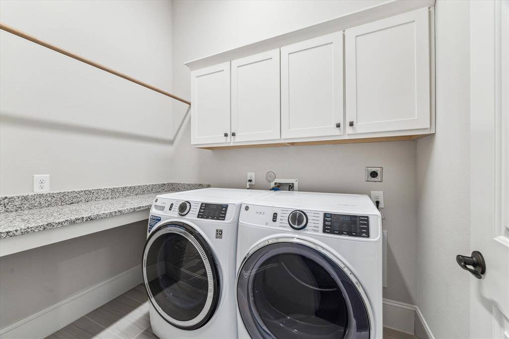 This first-floor laundry room offers ample storage with upper cabinetry, a spacious granite folding counter, and a built-in hanging rod for drying clothes. Neutral tones and durable tile flooring create a clean and functional space. Thoughtfully designed for efficiency, this laundry area provides everything needed to streamline daily chores.