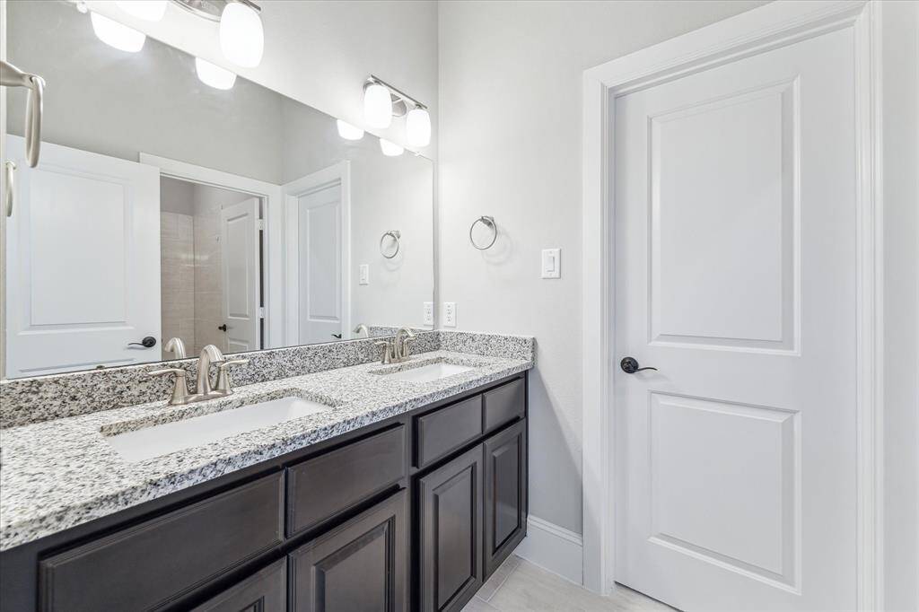 A spacious secondary bathroom with dual vanities, granite countertops, and brushed nickel fixtures for a sleek, modern look. Dark wood cabinetry provides generous storage, while the large mirror enhances the sense of space. Positioned between secondary bedrooms, this bathroom offers both convenience and privacy with a separate bathing area.