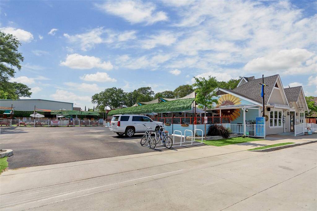 A bright and inviting shot of D'Alba, a beloved neighborhood eatery in Garden Oaks. The exterior features a charming mural of a sunflower on a blue backdrop, setting a cheerful and welcoming tone. The spacious patio, shaded with greenery-draped canopies, offers a relaxed outdoor dining experience. With bike racks out front and a convenient parking lot, it's a go-to spot for locals looking to enjoy fresh, seasonal fare in a laid-back setting.