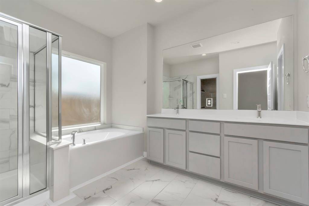Escape to a spa-like oasis in this primary bathroom. Enjoy a walk-in shower with tile surround, a separate garden tub with custom detailing, and an elegant vanity featuring light countertops, modern hardware, and beautiful vanity lights. Experience luxury and relaxation in every detail. Sample photo of completed home with similar floor plan. Actual colors and selections may vary.