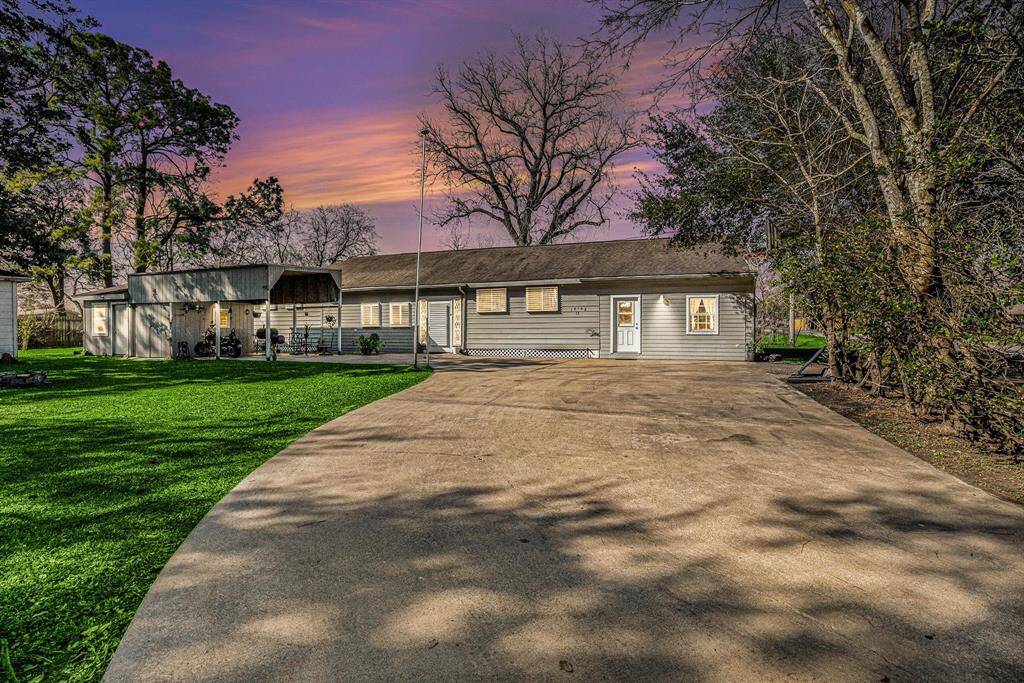 Charming single-story home with a spacious front yard, long driveway, and mature trees. The exterior features a cozy covered porch area and a serene setting under a vibrant sunset sky.