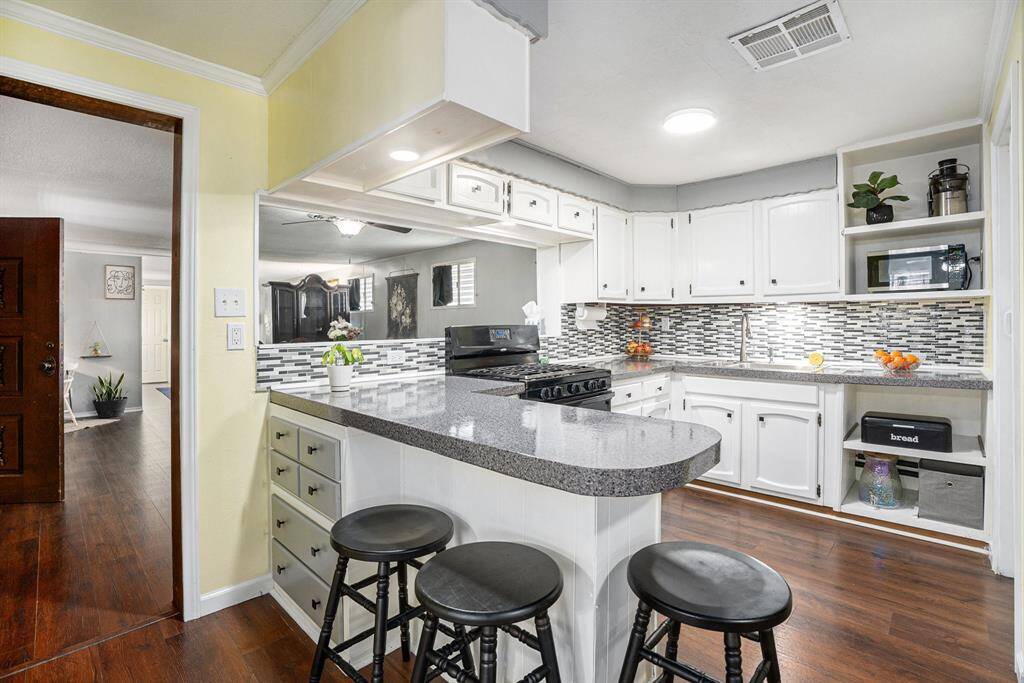 This photo showcases a bright, inviting kitchen with white cabinetry, a modern mosaic tile backsplash, and a sleek granite countertop. The space includes a cozy breakfast bar with stools, blending seamlessly into the adjacent living area, enhanced by rich hardwood floors and ample natural light.