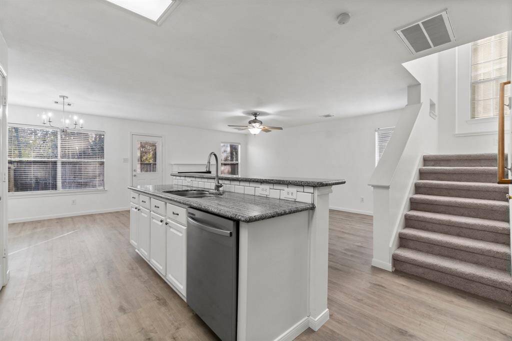 This photo showcases a modern, open-concept kitchen and living area with hardwood flooring. The kitchen features a granite island with a sink and stainless steel appliances, while nearby stairs lead to the upper level. Large windows provide ample natural light.