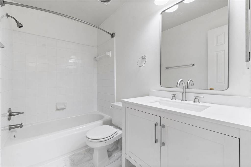 The secondary bathroom is bright and modern, featuring a white color scheme. It includes a bathtub with a shower, a sleek vanity with dual faucets, and a large mirror. The clean lines and neutral tones offer a fresh and inviting space.