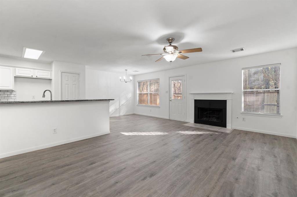 This bright living space features an open floor plan with wood-style flooring, a modern ceiling fan, and a cozy fireplace. The kitchen includes a breakfast bar and updated fixtures, adjacent to a dining area with natural light from large windows.