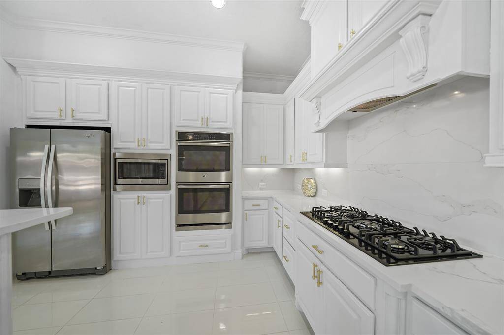 The kitchen features sleek white cabinets adorned with elegant gold handles, complemented by glossy white polished tiles and silver appliances , creating a bright, sophisticated atmosphere in a spacious elongated layout.